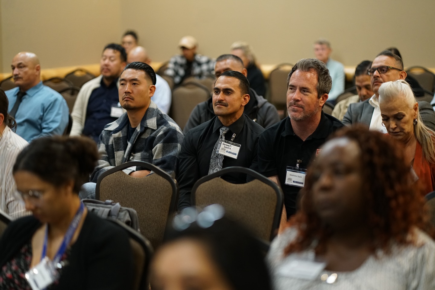 Diverse Professionals Attending Conference Session Diverse group of professionals, mostly men, seated in rows at a conference, many wearing name tags and looking forward.