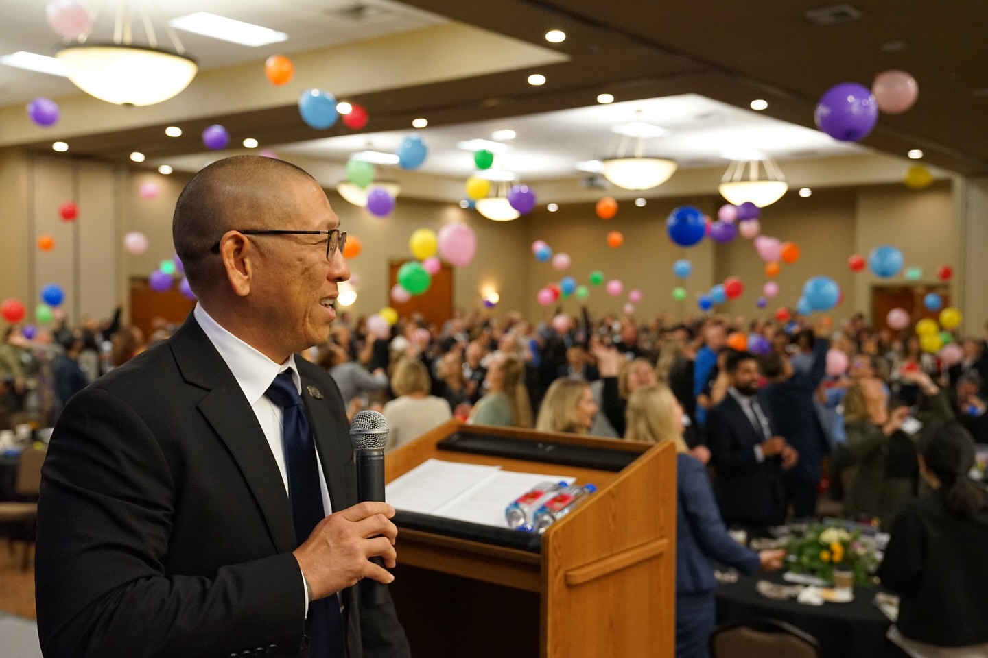 Speaker Addresses Audience at Festive Event with Balloons Man in suit and glasses speaking at a podium with a microphone, colorful balloons floating above a lively crowd in a large e…