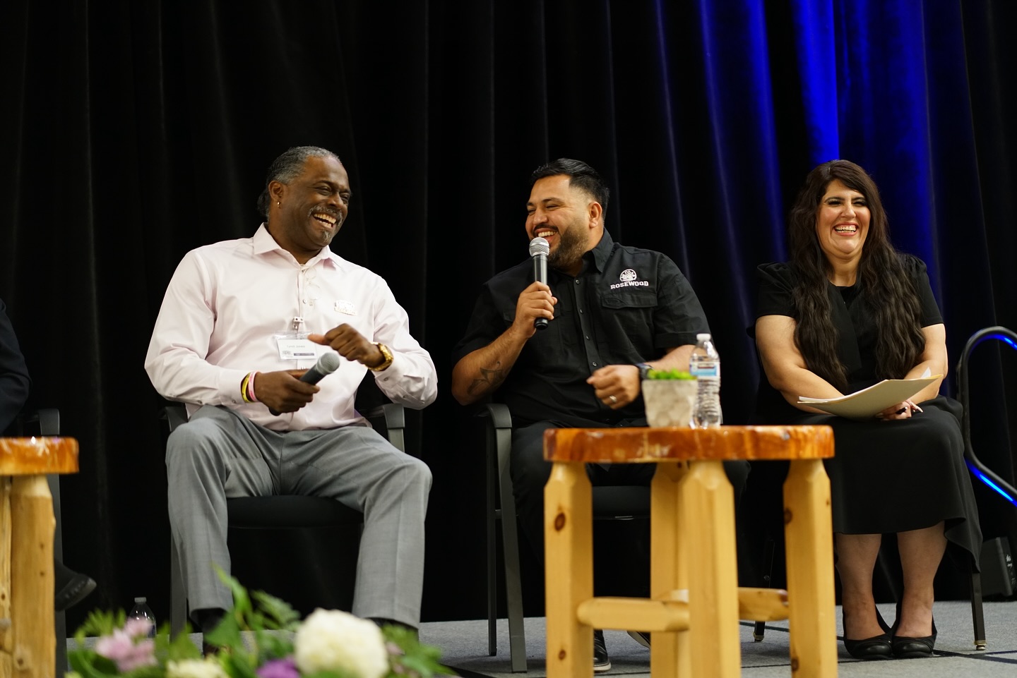 Engaging Panel Discussion at Professional Conference Event Three panelists, two men and one woman, smiling and laughing during a discussion on stage, with two holding microphones.