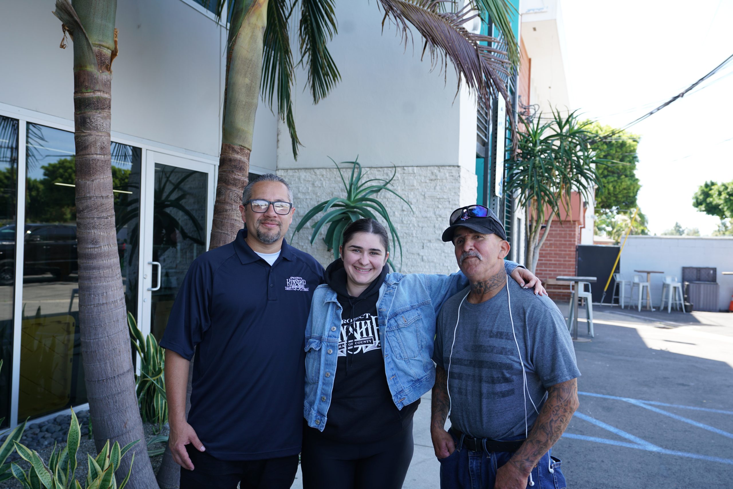 Three smiling individuals, two men and one woman, stand outdoors in front of a modern building with palm trees.