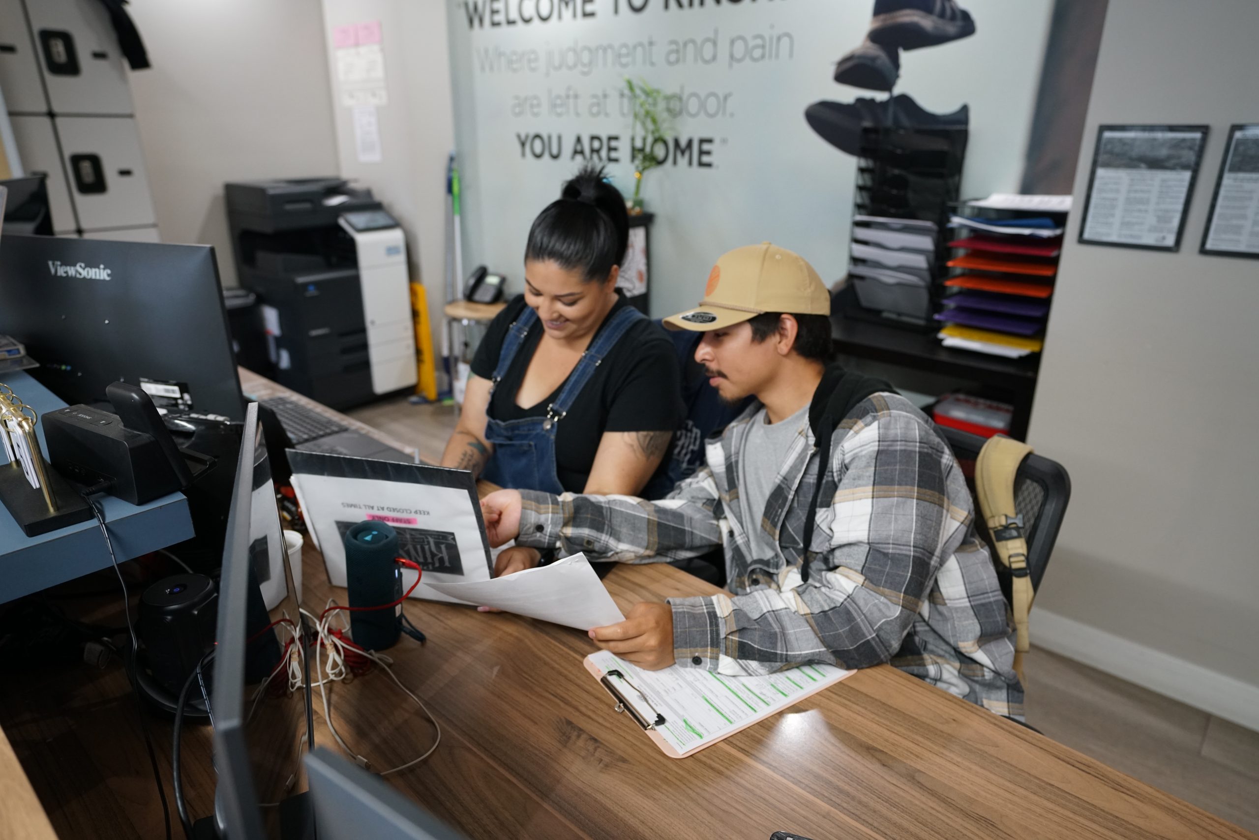 Two people, a smiling woman in blue overalls and a man in a plaid shirt, review documents together at a wooden office desk.