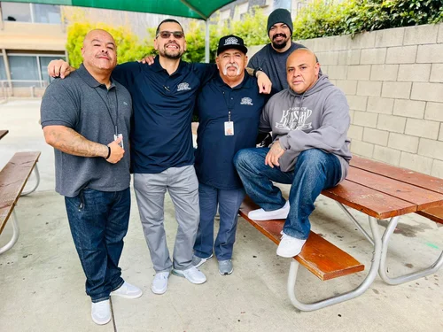 Adult Re-Entry Program Participants Smiling Together Five smiling adult men, casually dressed in polo shirts, hoodies, and jeans, pose together outdoors near a picnic table.