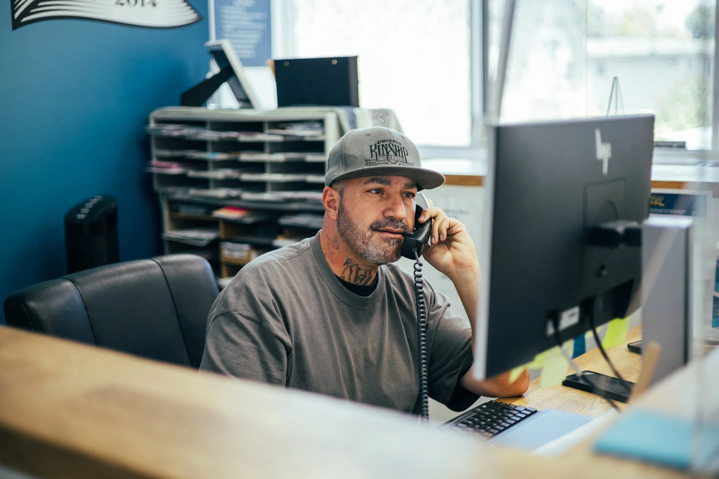 Man with beard and tattoos wearing a baseball cap, talking on a landline phone at an office desk with a computer.