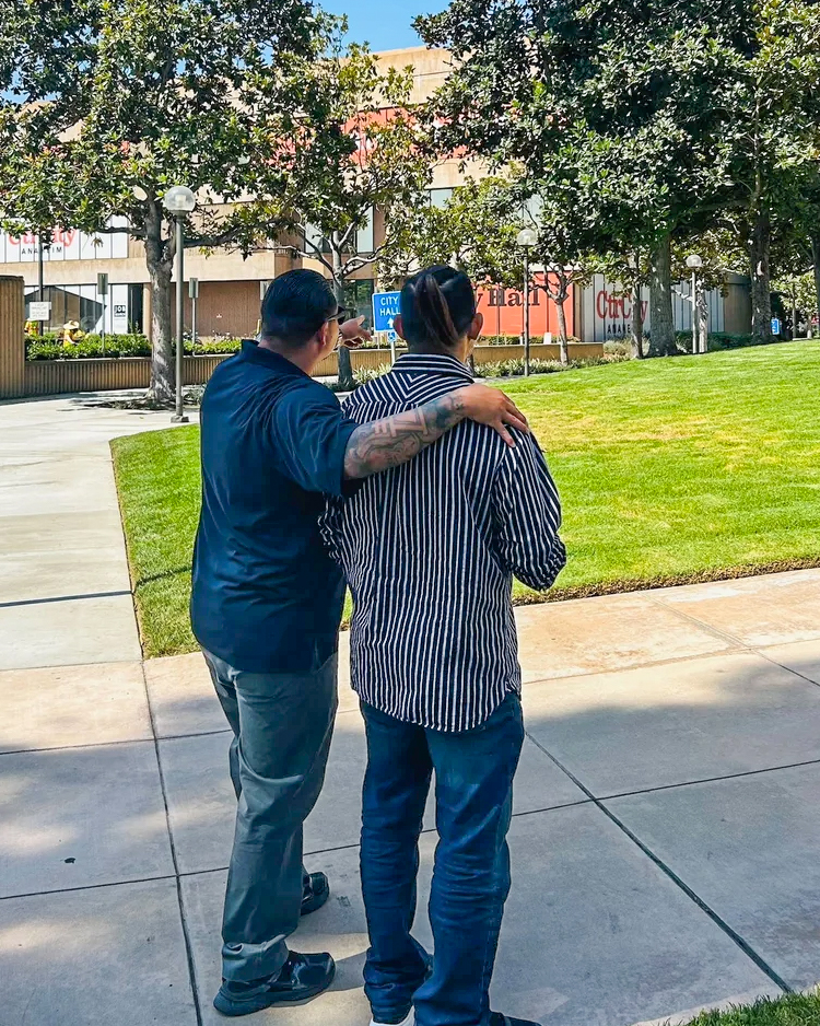 Mentorship at Anaheim City Hall for Corrections Cohort Two men from behind, one with arm around the other, pointing towards Anaheim City Hall. Sunny outdoor scene with trees and g…