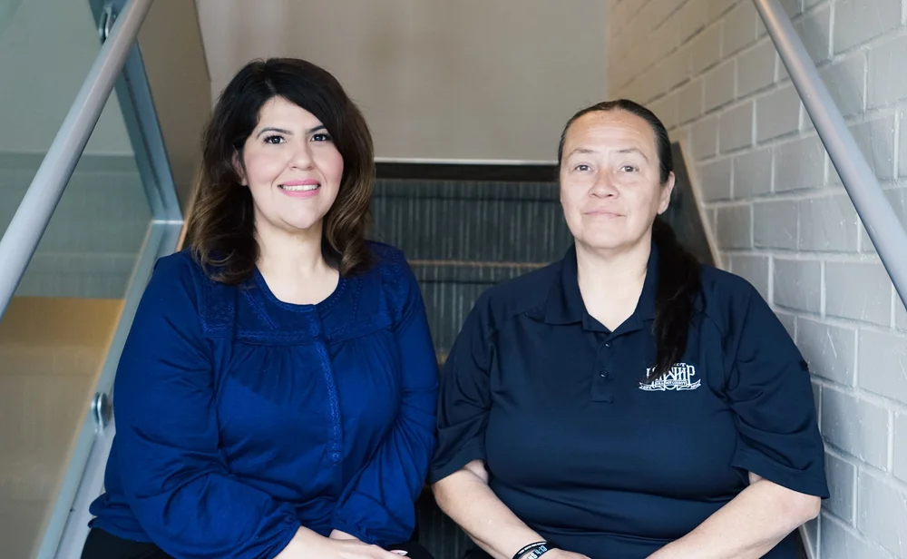 Dedicated Enhanced Care Management Team Members Two women, one with dark wavy hair and a blue blouse, the other with dark hair in a ponytail and a navy polo, sit on stairs.