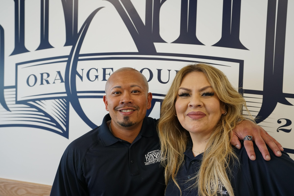 Project Kinship Orange County Leadership Team Portrait Man with goatee and woman with long blonde hair smiling, wearing Project Kinship polo shirts, in front of an "ORANGE COUNTY"…
