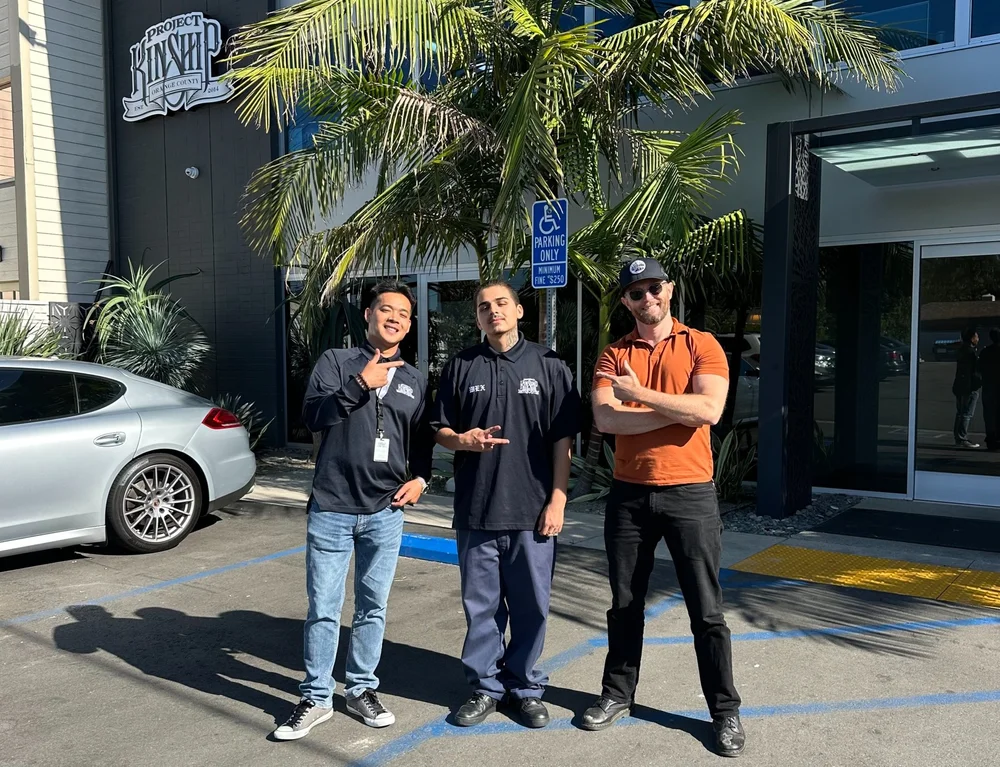 Project Kinship Team Members Outside Office Building Three men, smiling and gesturing, stand outside the Project Kinship building with a silver Porsche and palm tree in the back…