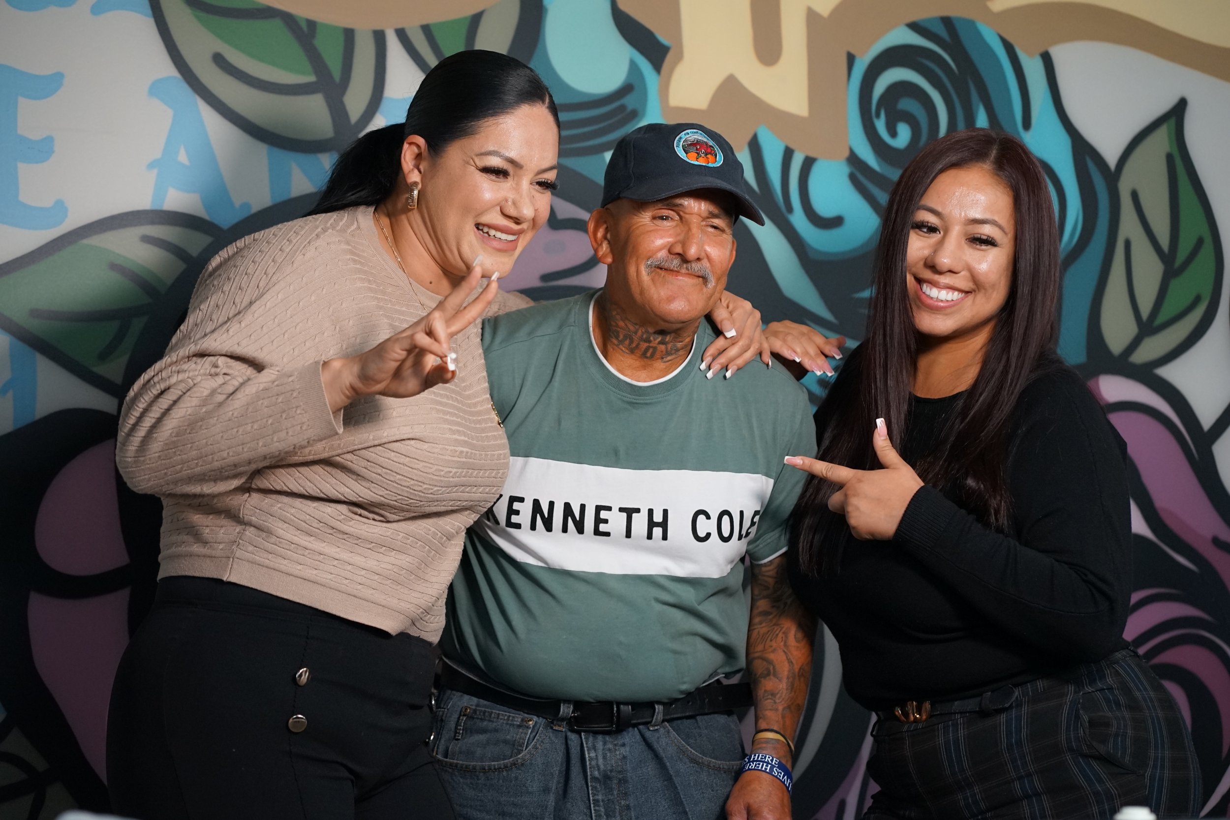 Three smiling people, two women and one man, posing happily in front of a colorful mural with abstract art.