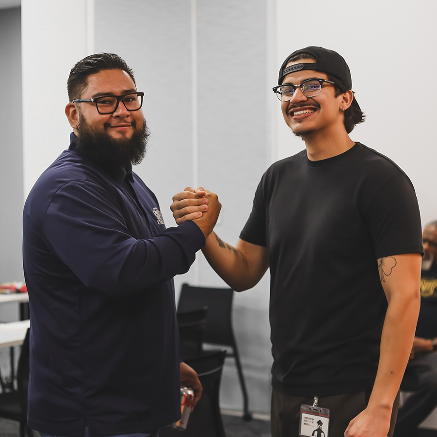 Friendly Handshake Between Two Men, Smiling Two men, one bearded in glasses and a navy jacket, the other in a black t-shirt and backward cap, shake hands and smile.