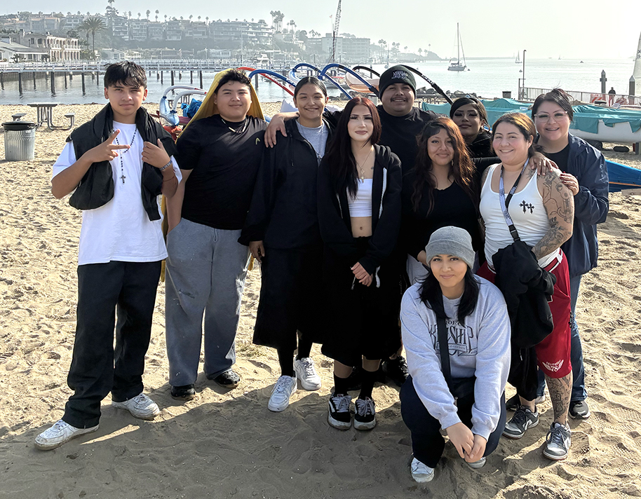 Youth Program Group Enjoys Sunny Beach Day Ten young people, diverse in appearance, pose casually on a sunny sandy beach with a harbor, boats, and distant buildings.