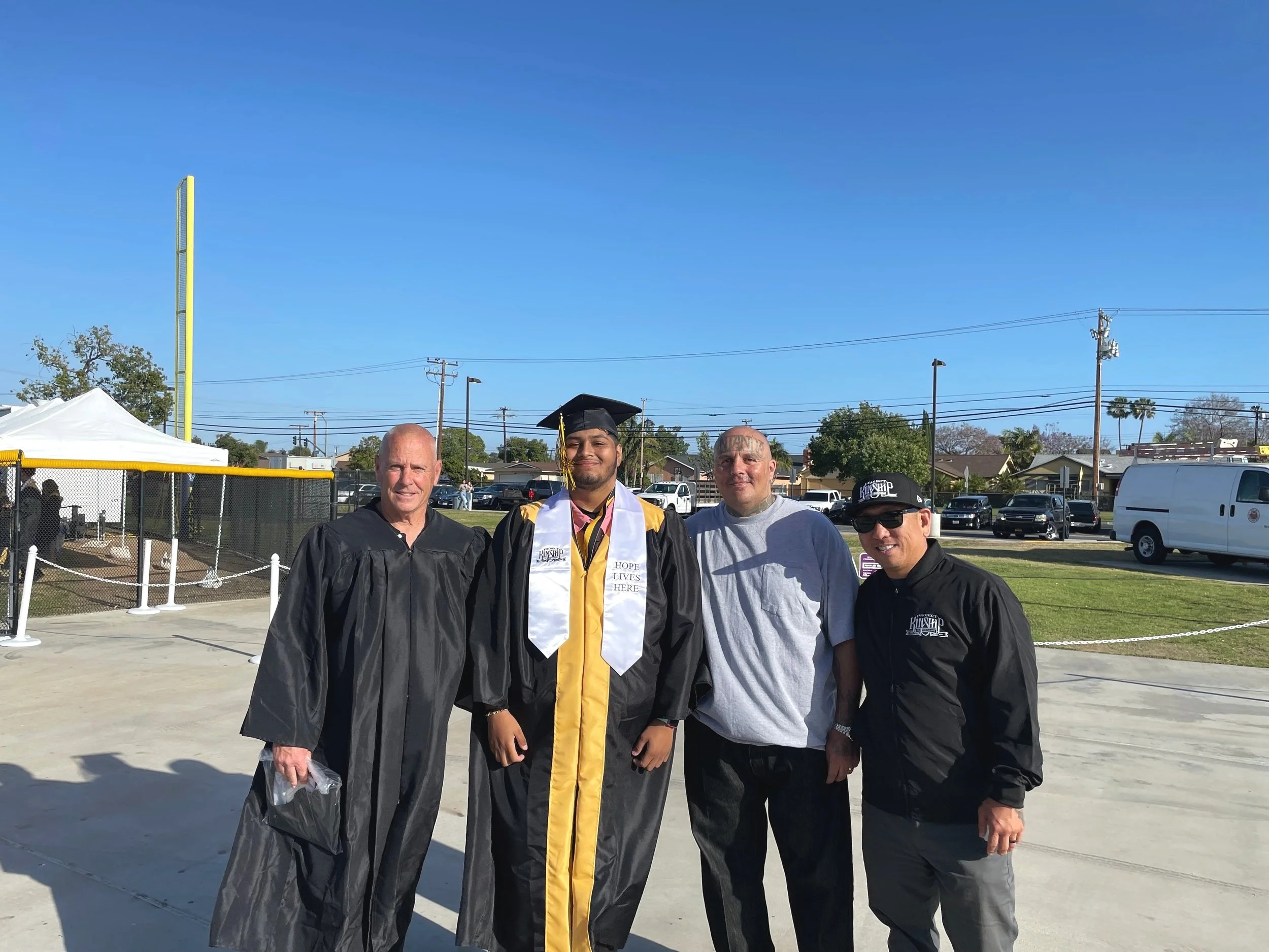 Four men, including a graduate in a cap and gown, pose outdoors on a sunny day near a baseball field.