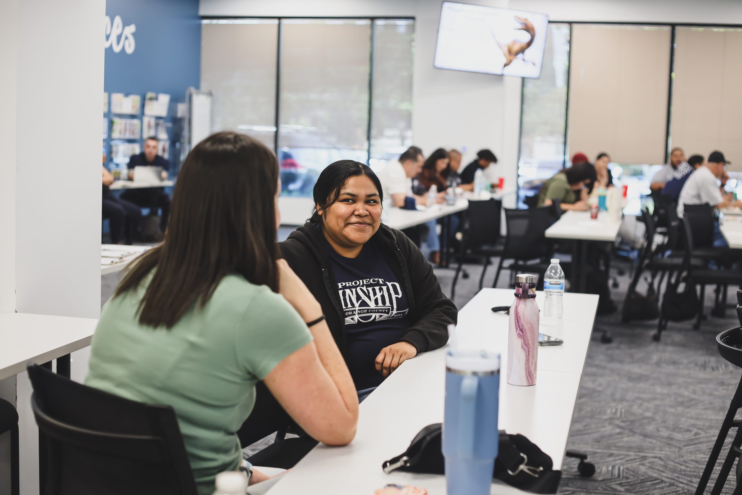 Two women converse at a workshop table, one smiling at the camera in a Project Kinship shirt, surrounded by other participan…