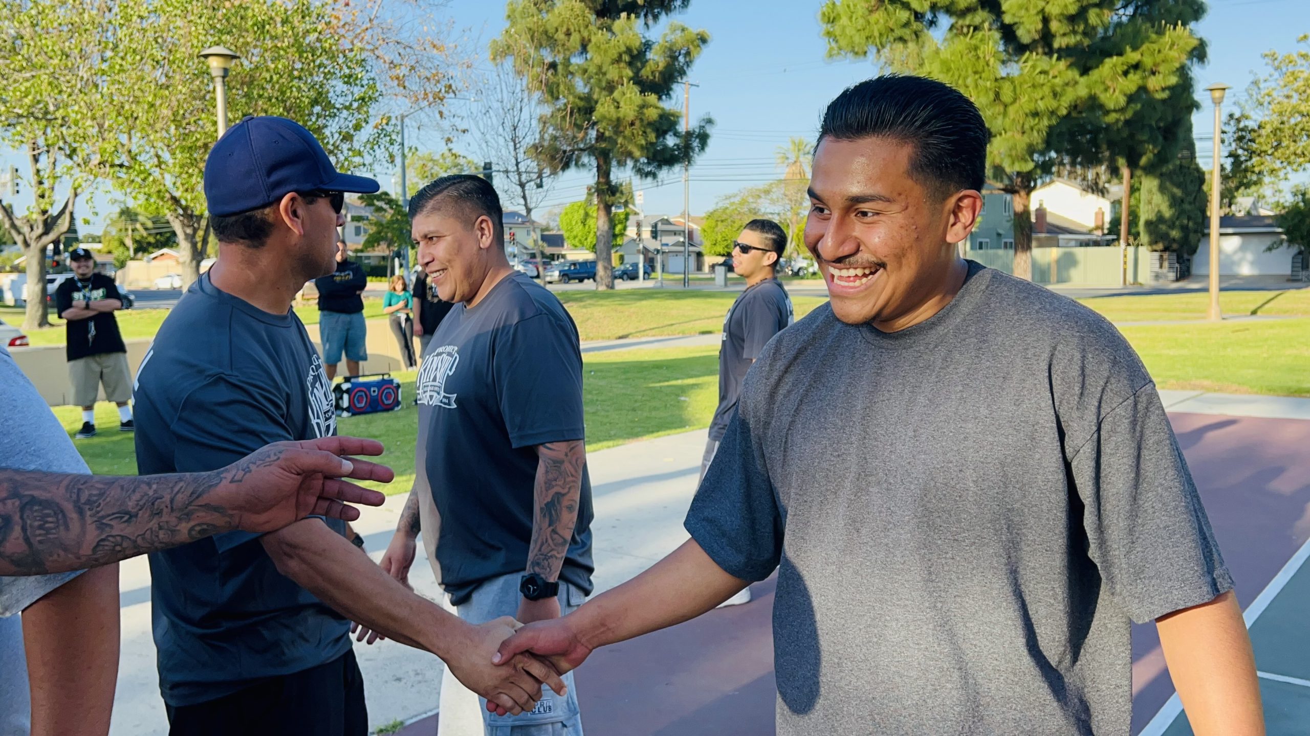 Two men warmly shaking hands and smiling in a sunny park, with other people and trees in the background.