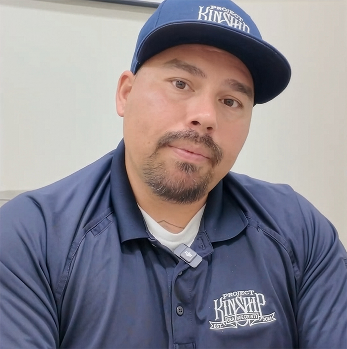 A man with a goatee and mustache, wearing a navy blue 'Project Kinship' baseball cap and polo shirt, looking directly at the…