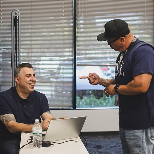 Engaged Learning and Collaboration in Adult Programs Two men interact in a classroom: one seated, laughing at a laptop; the other standing, pointing playfully.