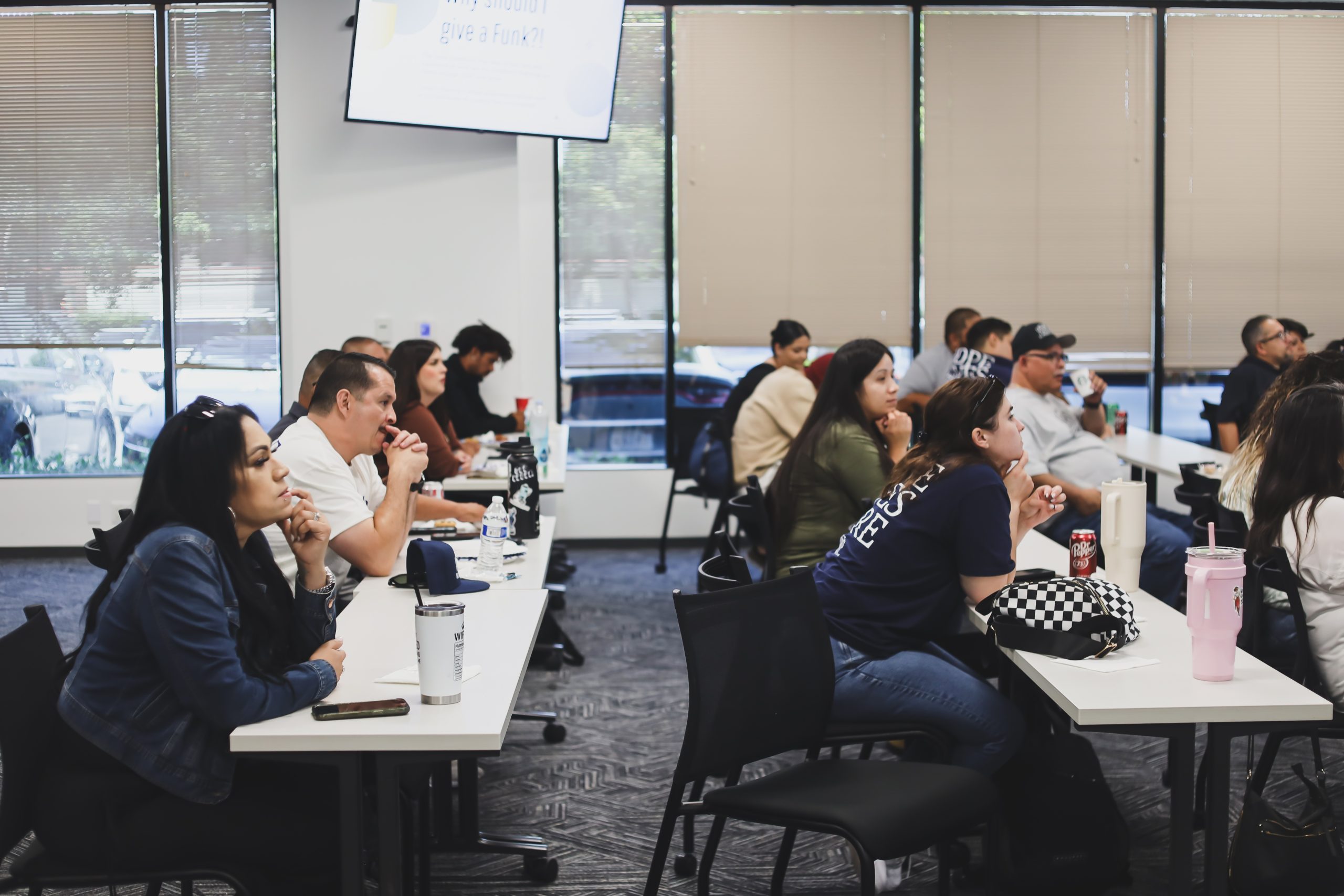 Diverse group of adults attending a presentation in a bright room, seated at tables with drinks and personal items.