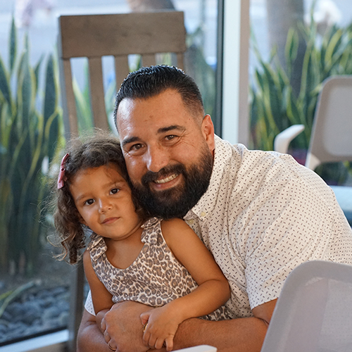 Father and Daughter Share a Happy Moment A smiling man with a beard holds a young girl with curly hair, who rests her head on his shoulder, both looking at the camer…