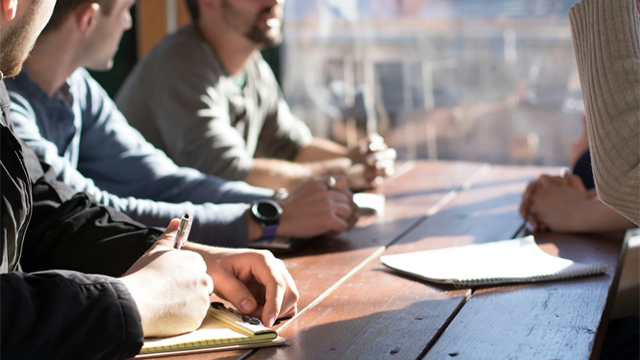 Four men at a wooden table, one actively writing notes on a yellow pad during a focused group discussion.