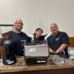 Three smiling baristas, two men and one woman, stand behind a mobile coffee bar with a grinder, espresso machine, and sink.