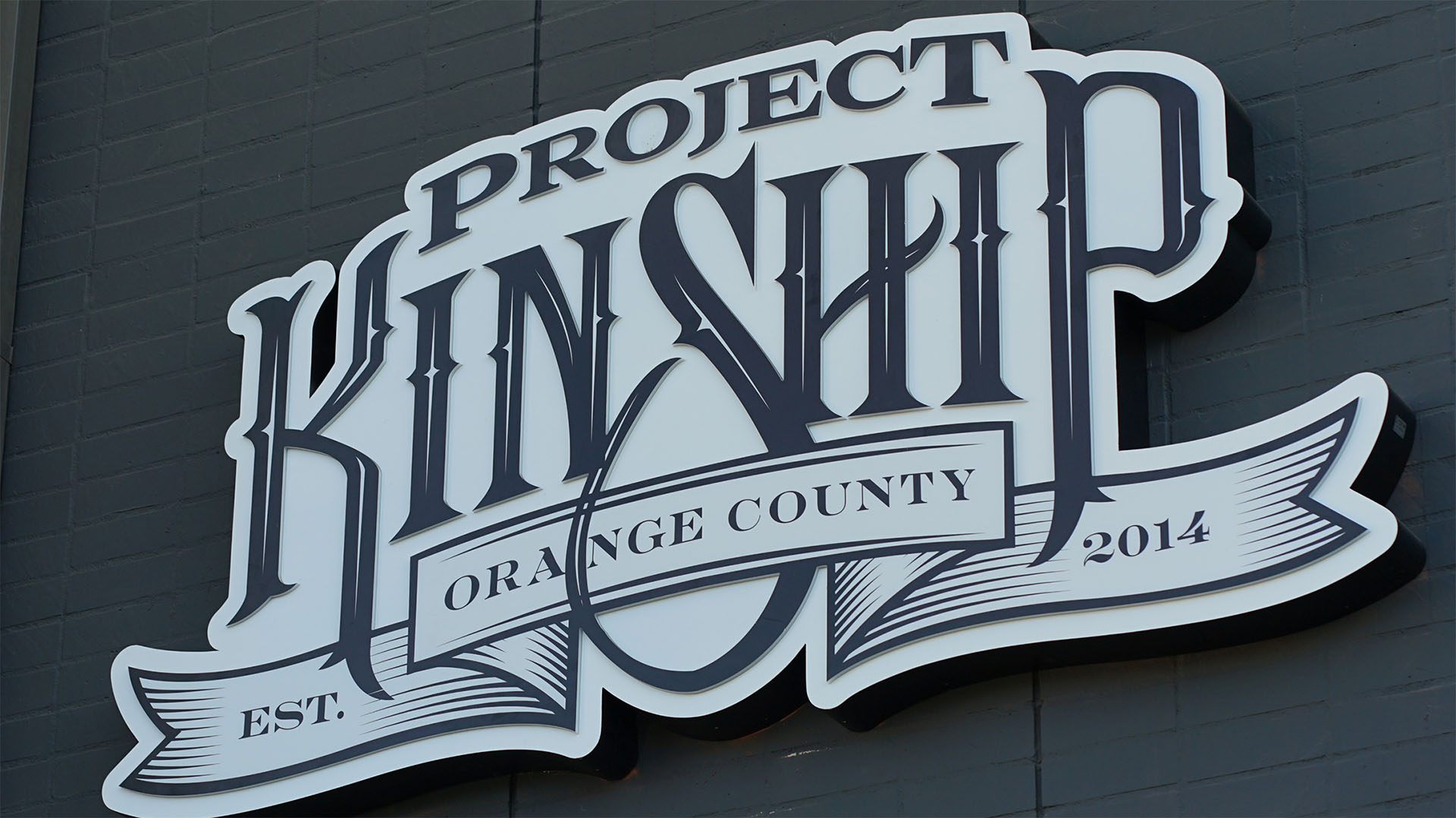 This eye-catching sign features "PROJECT KINSHIP" in a prominent, ornate dark gray font against a white background, creating a striking 3D effect. A banner across the center reads "ORANGE COUNTY," flanked by "EST." and "2014," indicating its founding. The sign is mounted on a dark gray brick wall, showcasing its craftsmanship and brand identity.