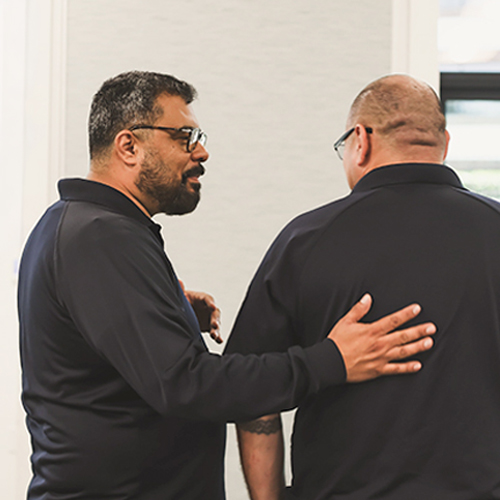 Supportive Colleagues Connecting at a Professional Event Man with beard and glasses smiles at another man, placing a supportive hand on his back. Both wear dark shirts.