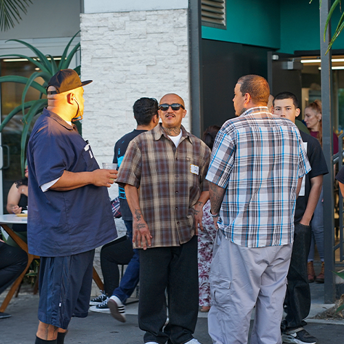 Men in Plaid Shirts Conversing at Outdoor Gathering Three men, two in plaid shirts and one in a dark blue shirt, conversing outdoors; one man wears sunglasses and neck tattoos.