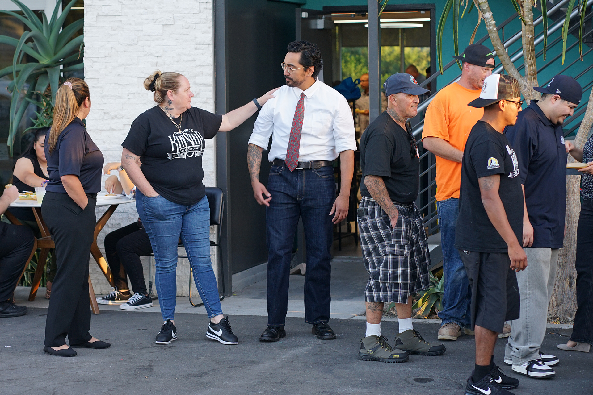 Diverse group of people gathered outdoors, a woman touches a man's shoulder, near a building entrance with plants.