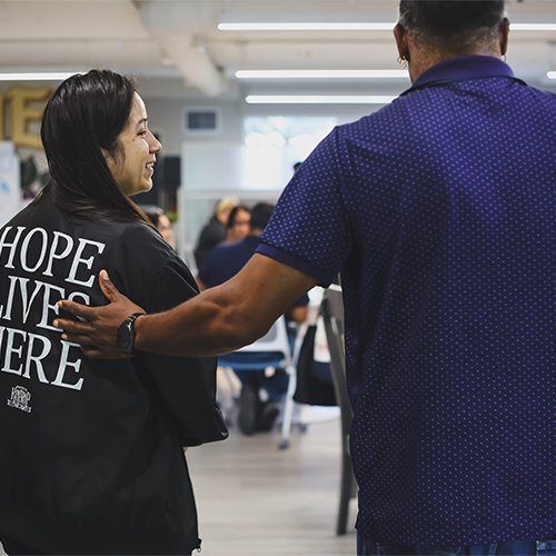 Supportive Interaction at Project Kinship A man's hand rests supportively on the shoulder of a smiling woman wearing a black shirt that reads 'HOPE LIVES HERE'.