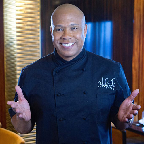 Chef Jeff Henderson Welcomes Public Safety Conference Attendees Smiling bald man in black chef's coat with open hands, gesturing welcomingly to an audience.