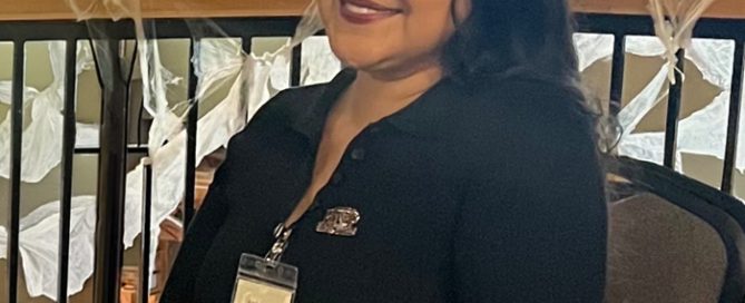 April, a smiling woman with dark hair and a black collared shirt, wearing an ID badge, sits at a table with hands clasped.
