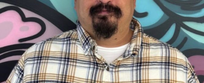 Bald man with a dark goatee wearing a white, brown, and blue plaid shirt, smiling against a vibrant, abstract mural.