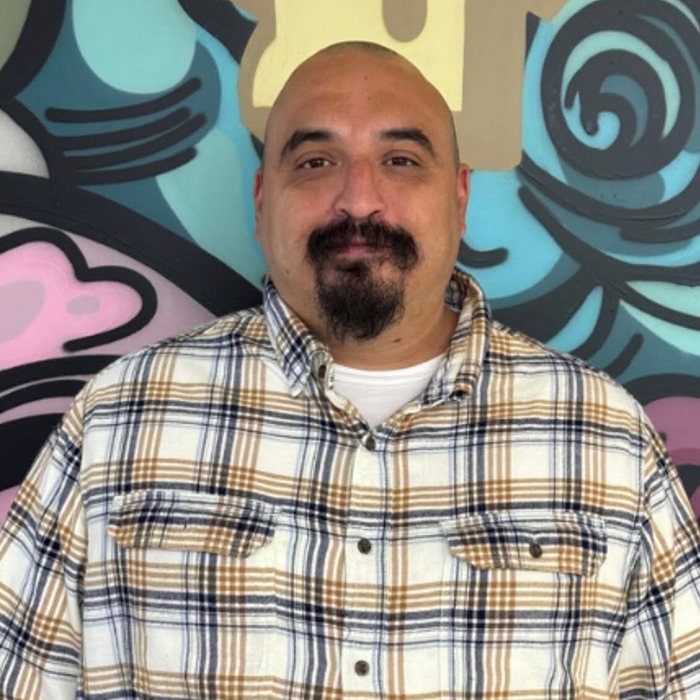 Bald man with a dark goatee wearing a white, brown, and blue plaid shirt, smiling against a vibrant, abstract mural.