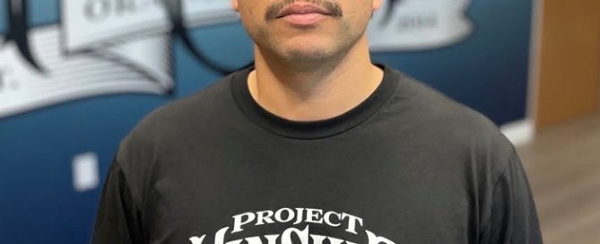 Bald man, Arturo, with glasses and mustache, wearing a black "Project Kinship Orange County" shirt, standing before a blue m…