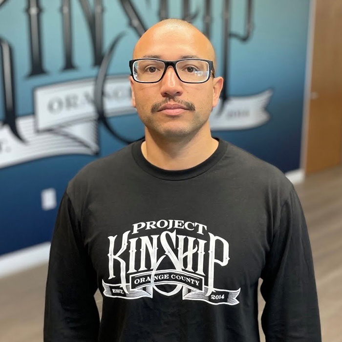 Bald man, Arturo, with glasses and mustache, wearing a black "Project Kinship Orange County" shirt, standing before a blue m…