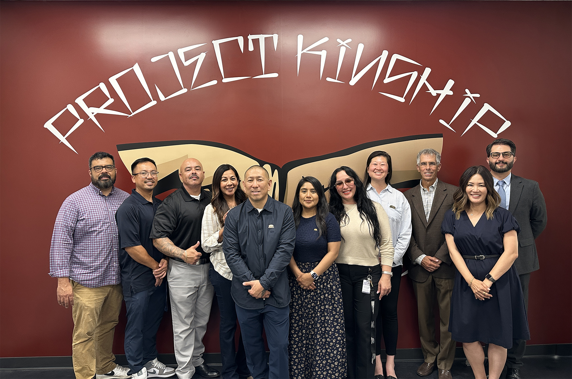 Eleven diverse individuals, men and women, smiling and standing together in front of a red wall with 'PROJECT KINSHIP' in wh…
