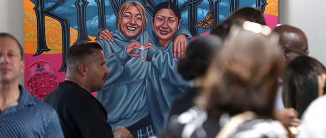 A vibrant mural depicts two smiling women in blue hoodies forming a heart, with 'KINSHIP' text above, as people gather.