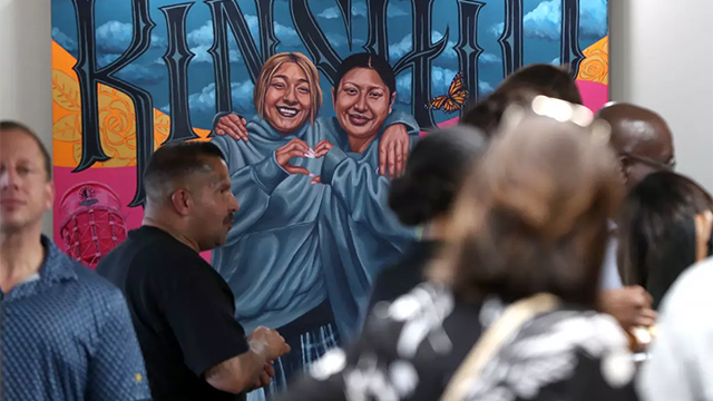 A vibrant mural depicts two smiling women in blue hoodies forming a heart, with 'KINSHIP' text above, as people gather.
