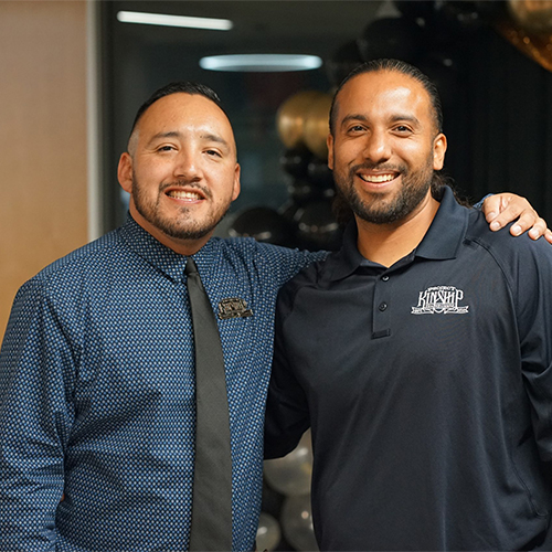 Project Kinship Team Members Smiling Together Two smiling men, one in a blue patterned shirt and tie, the other in a navy polo with a 'Kinship' logo, stand together.