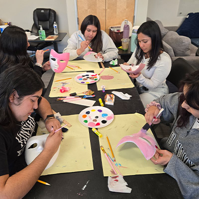 Young Women Painting Masks at Creative Art Workshop Five young women painting decorative masks with colorful paints and brushes at a long table during an art workshop.