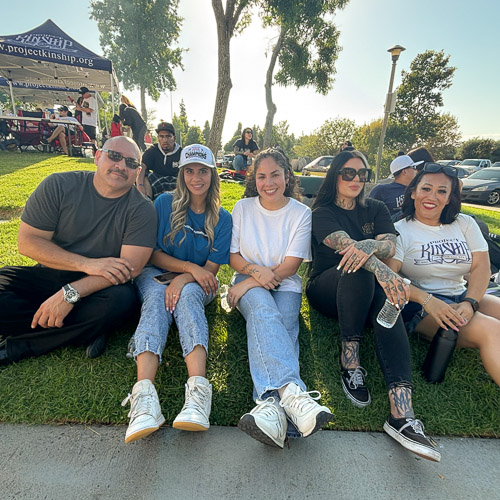 Project Kinship Community Event Gathering on Grass Five adults, three women and two men, sit on green grass, smiling at an outdoor community event with a 'Project Kinship' ten…