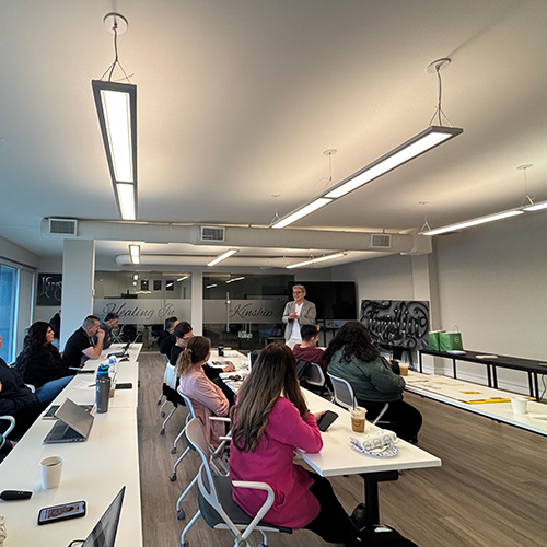 Professional Presentation at Healing Institute with Engaged Audience Speaker addresses a diverse group in a modern training room with frosted glass partitions, linear lights, and long white tab…